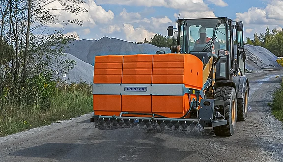 Ein Radlader auf einem unbefestigten Weg, der mit einem Tanksystem ausgestattet ist. Im Hintergrund sind große Materialhaufen zu sehen, während Bäume und ein blauer Himmel die Szene umrahmen.