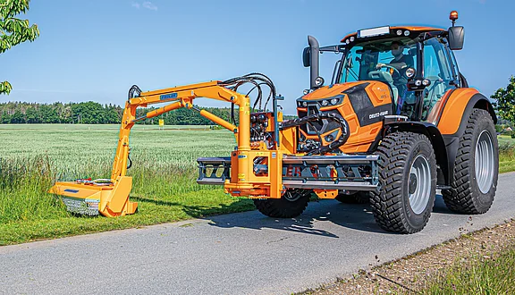 Ein orangefarbener Traktor steht am Straßenrand, ausgestattet mit einem großen Frontausleger mit Mähkopf. Das Gerät ist zum Mähen von Gras oder Unkraut ausgelegt und befindet sich in der Position, um die Arbeit zu verrichten. Im Hintergrund sind grüne Felder und Bäume sichtbar.