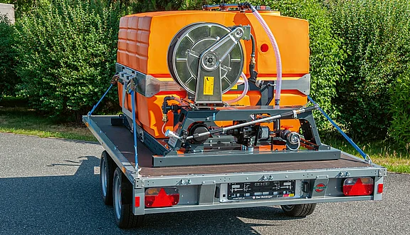 Das Bild zeigt einen orangefarbenen Wassertank auf einem Anhänger. Der Tank ist mit einer Pumpe und anderen Anbauteilen ausgestattet und für den Transport auf einer befestigten Straße vorgesehen. Im Hintergrund sind grüne Sträucher sichtbar.