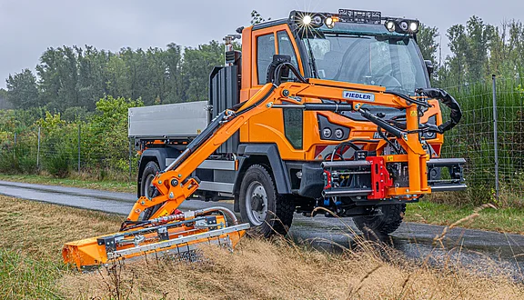 Ein M41 mit einem seitlichen ausgeklappten Frontausleger mit Mähwerk steht auf einer nassen Straße und mäht den Straßenrandstreifen. Der Multicar hat eine Ladefläche. Im Hintergrund sind Bäume und eine Zäunung zu sehen.