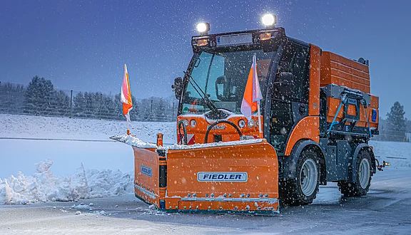 Ein orangefarbener Schneepflug steht in einer verschneiten Landschaft. Der Pflug ist mit Warnflaggen ausgestattet und die Umgebung ist von leichten Schneeflocken bedeckt. Die Dämmerung sorgt für eine kalte, winterliche Atmosphäre.