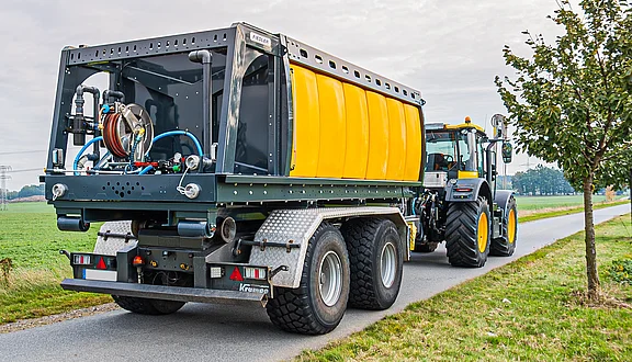 Ein Traktor zieht einen großen, gelben Tankanhänger. Der Anhänger hat eine komplexe technische Ausstattung. Die Umgebung ist ländlich, mit Feldern im Hintergrund und Bäumen entlang der Straße. An der Front hat der Traktor einen Gießarm montiert.
