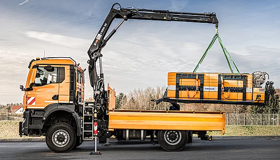 Ein orangefarbener MAN mit einem Kran hebt ein großes, orangefarbenes Tanksystem an, um es auf seine Ladefläche zu positionieren. Der Hintergrund zeigt eine klare Himmelspartie und eine bewaldete Fläche.