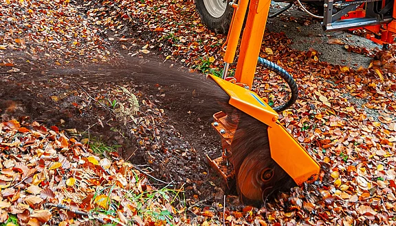 Ein orangefarbener Grabenfräse an einem Frontausleger, fräst einen Graben für den Wasserablauf in die Erde. Umgeben von bunten Herbstblättern, zeigt die Maschine ihre Anwendung in der Landschafts- oder Gartenerstellung.