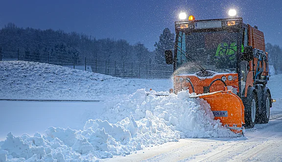 Ein orangefarbener Schneepflug entfernt Schnee von einer Straße während einer Schneefallnacht. Im Hintergrund sind verschneite Bäume und eine grüne Umzäunung zu erkennen. Die Beleuchtung des Schneepflugs sorgt für Sichtbarkeit im winterlichen Wetter.