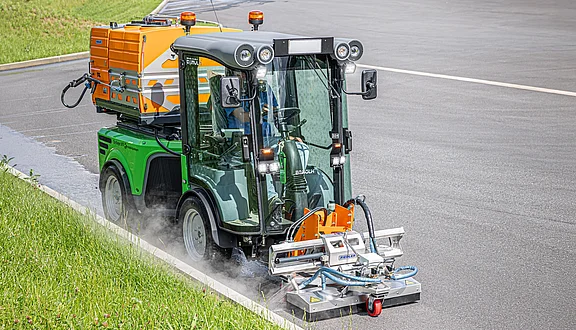 Ein grüner Reinigungsfahrzeug mit einer Heißwasseranlage fährt auf einer Straße. Es hat einen Unkrautvernichter montiert und wird für die Pflege der Straßenfläche eingesetzt. Dampf tritt aus, was zur Bekämpfung von Unkraut angewendet wird.