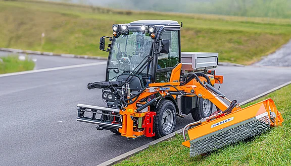 Ein kleines Kommunalfahrzeug steht an einer Straße, ausgestattet mit einem Frontausleger und Mähkopf. Der Mähkopf ist auf einem grünen Hang positioniert. Der Hintergrund zeigt eine bewachsene Landschaft.