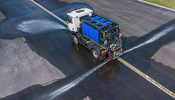 Ein MAN sprüht Wasser mit Schwemmdüsen auf eine asphaltierte Straße um sie zu Reinigen. Der Wagen ist mit einem blauen Wassertank ausgestattet und hinterlässt feuchte Spuren auf der Fahrbahn. Die Aufnahme zeigt die Perspektive von oben.